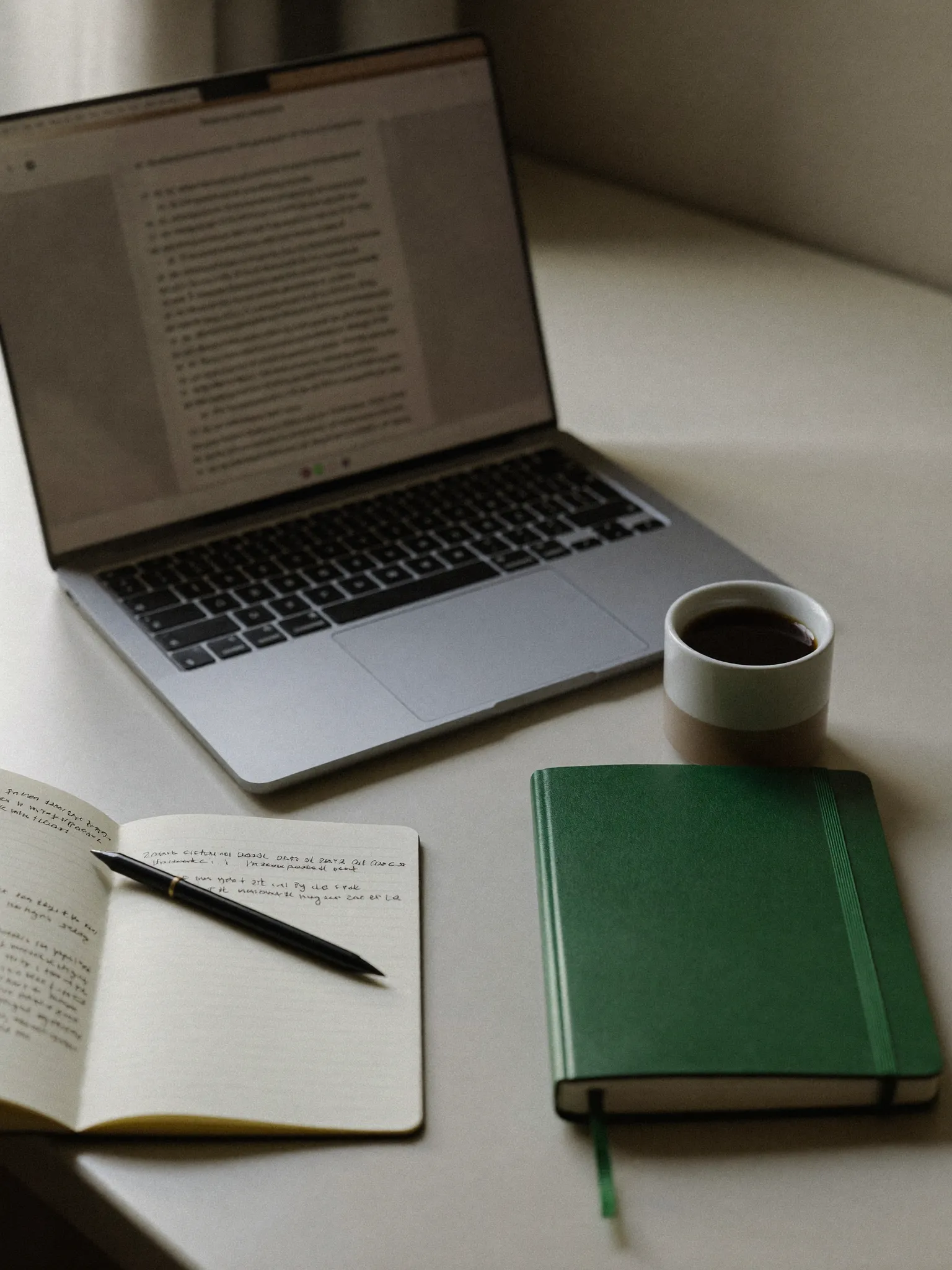 A desk with an open laptop, a handwritten testing notebook, coffee, and a green notebook